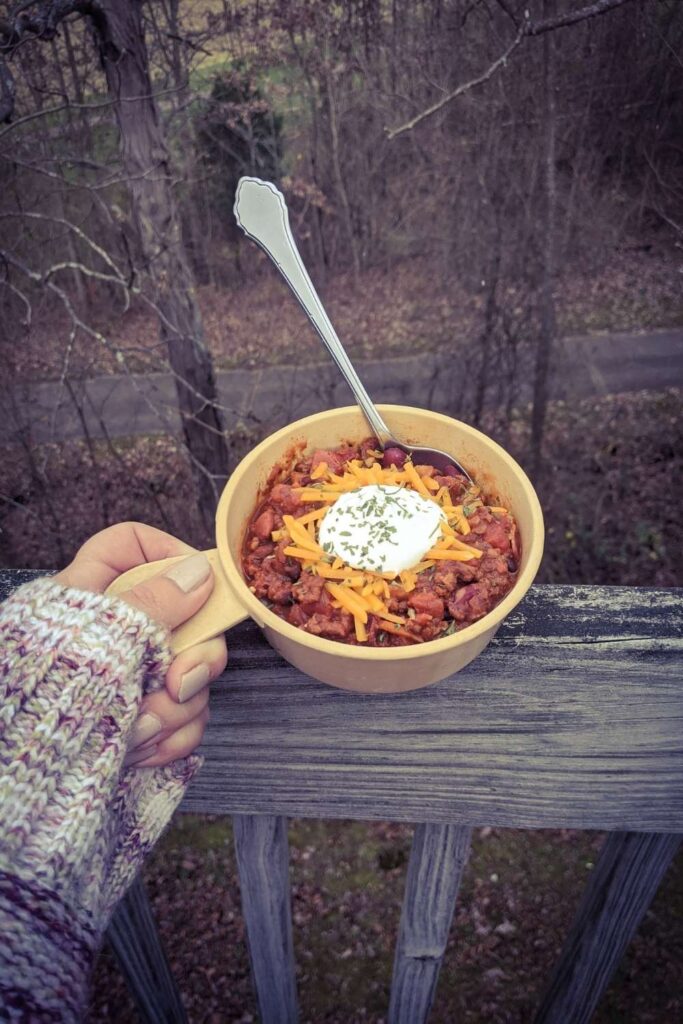 Haute Chili. Yummm. 8 Photograph of hand holding a beige bowl with a thumb-sized handle over wooden fence overlooking fallen autumn leaves and bare trees.