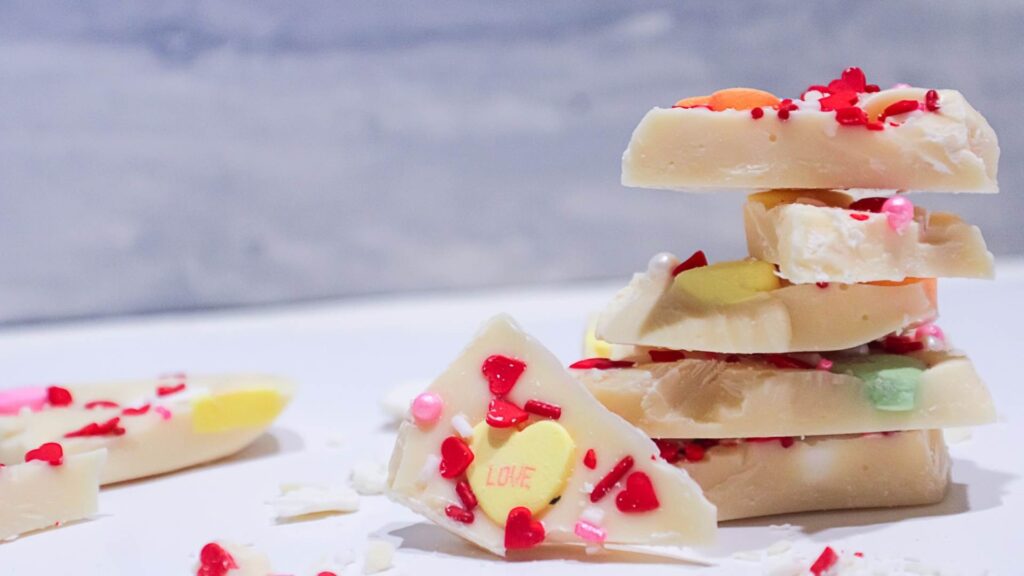 White Chocolate Sweethearts Bark made with love 8 White Chocolate Sweethearts Bark. 5 Chunks of bark stacked with one chunk facing front leaning on stack showing yellow conversation heart that says, love, and red heart sprinkles. with some shaved chocolate and sprinkles around it on a white table with a gray background.