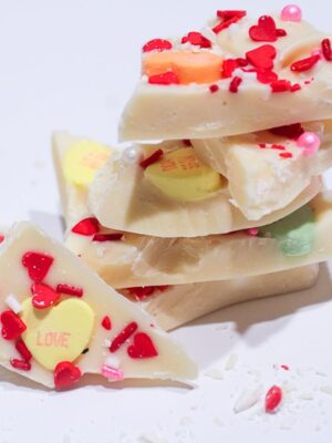 White Chocolate Sweethearts Bark. 5 Chunks of bark stacked with one chunk facing front leaning on stack showing yellow conversation heart that says, love, and red heart sprinkles. with some shaved chocolate and sprinkles around it on a white table with a gray background.