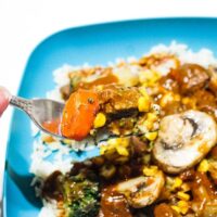 Quick and Easy Steak Glop Recipe Card Image featuring a fork with steak and carrot on it, and a blurred background of a teal bowl with white rice piled with california style veggies, mushrooms, corn, steak, and tons of gravy.