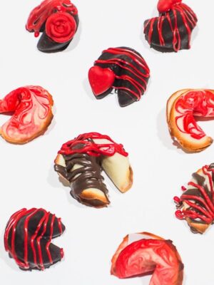 Photo of fortune cookies covered in drippy gooey milk chocolate and red chocolate foe shizzle drizzles on a cooling rack on a white background.