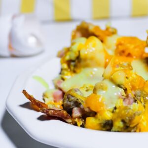 photo of eggs with sausage, bacon, cheddar cheese and hollandaise sauce on a white plate, on a white counter, with egg shells sparkling in the background in front of a yellow and white towel