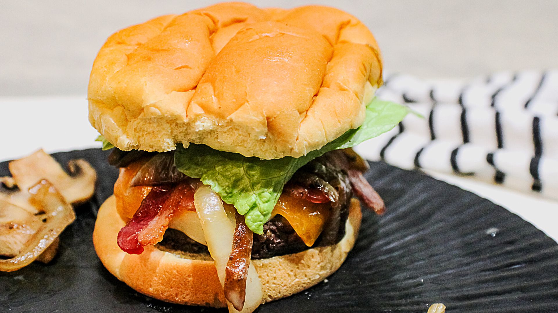 Cheyenne Cowboy Burger (Authentic Wyoming Recipe) 4 cheyenne cowboy burger on a black plate on gray background with some mushrooms on the side. Bacon, cheese, lettuce and burger are visible underneath a bun